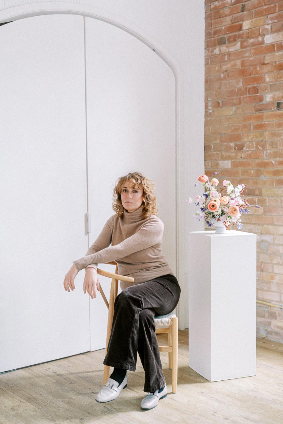A woman in a beige turtleneck and brown corduroy pants sitting on a wooden chair, next to a pedestal with a colorful floral arrangement, set against a brick wall interior backdrop.