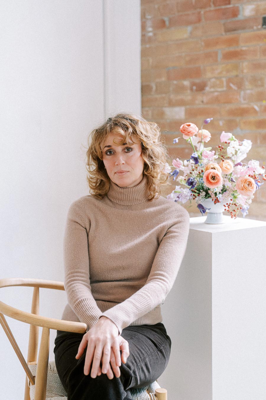 A person sitting on a wooden chair in a bright room, wearing a beige turtleneck sweater and dark pants, with a colorful flower arrangement on a pedestal nearby against a brick wall background.