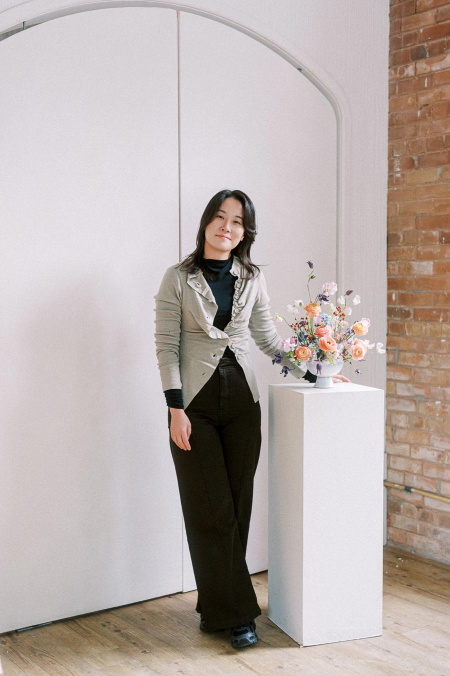 A person standing next to a white pedestal with a colorful flower arrangement, in a room with a white door and brick wall.