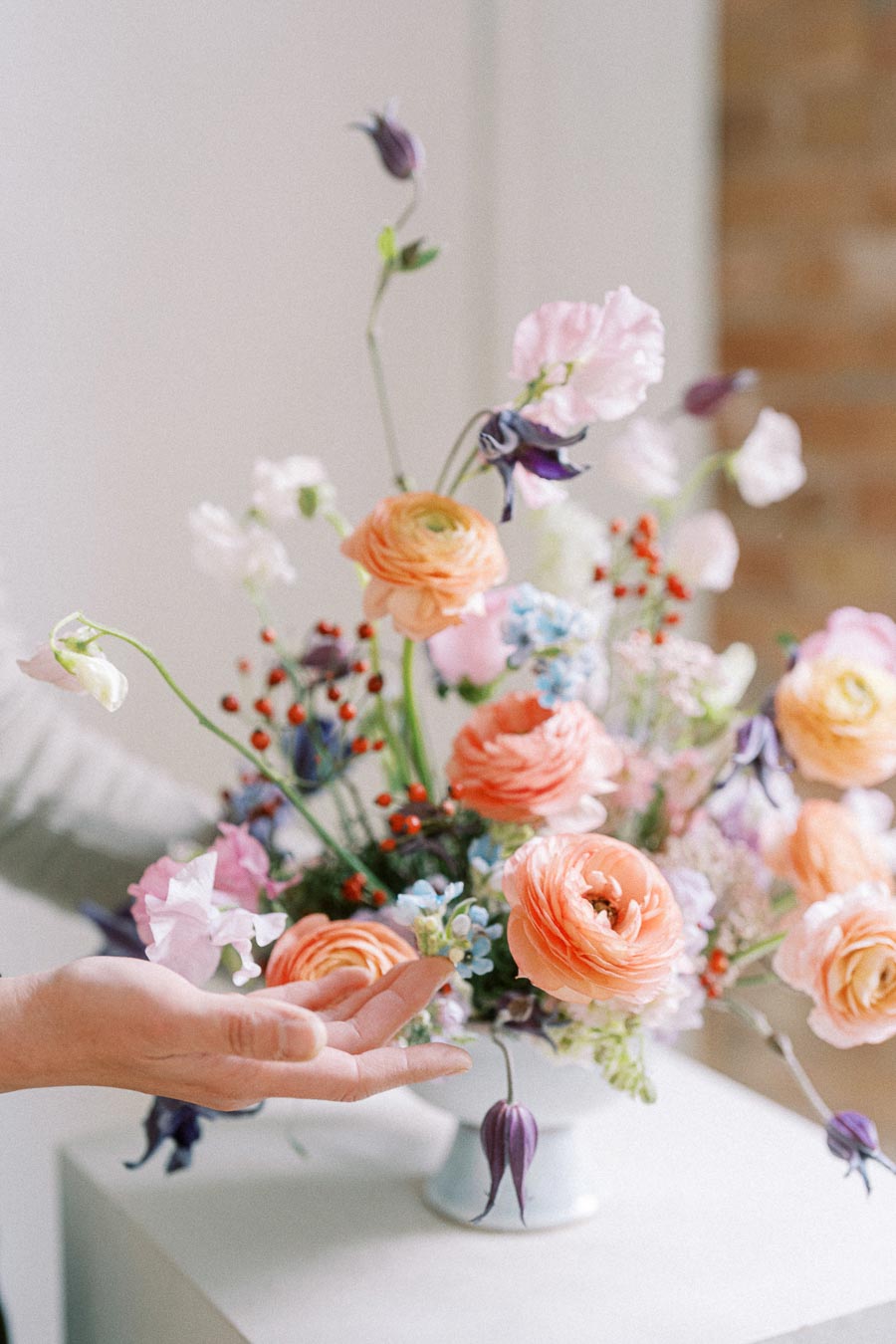 A vibrant bouquet of peach ranunculus flowers, purple sweet peas, and red berries in a white vase displayed on a table, with a hand arranging the floral arrangement for decoration.