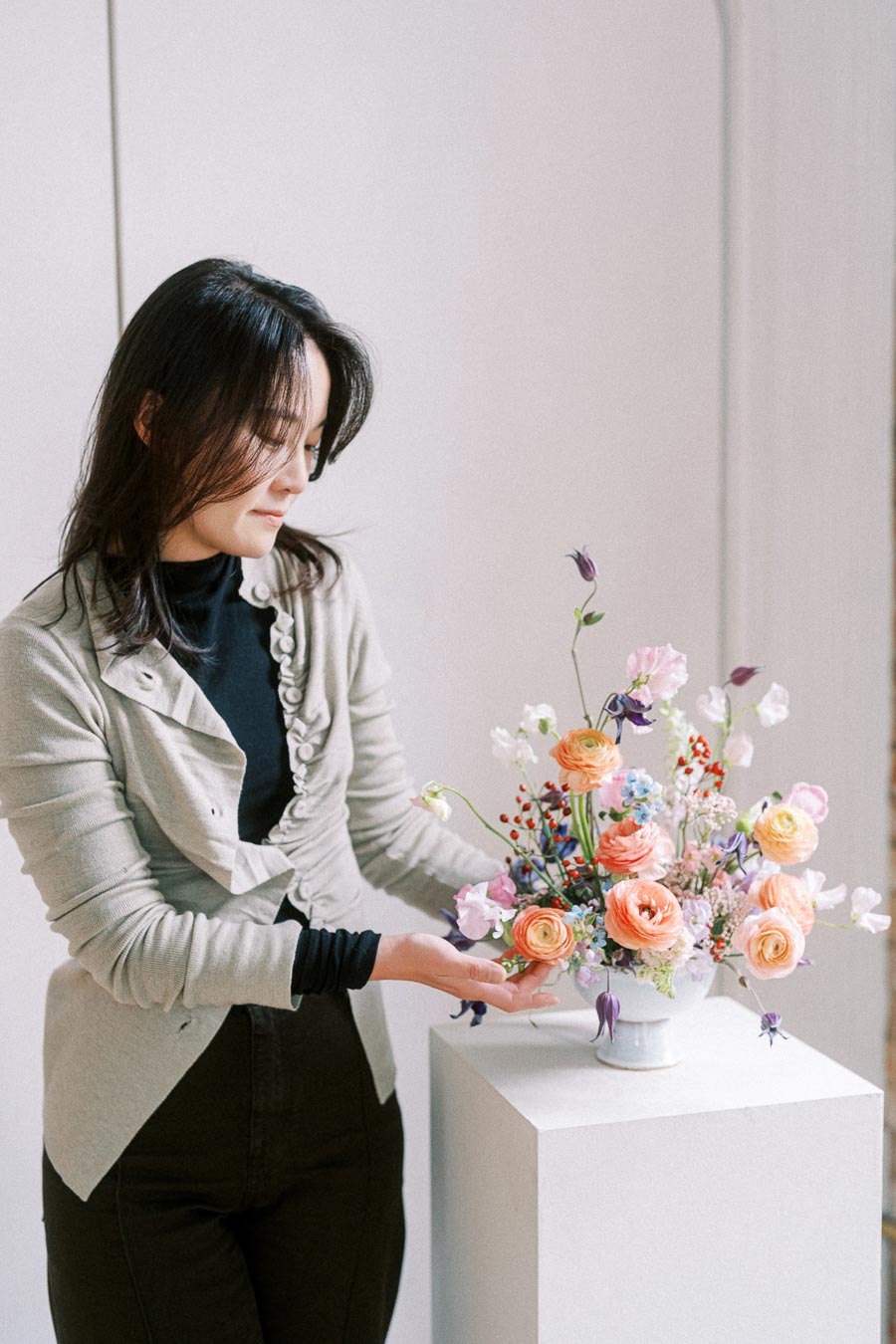 A person arranging a vibrant floral bouquet with pastel roses, wildflowers, and greenery in a white vase on a pedestal, showcasing a delicate and artistic floral design.
