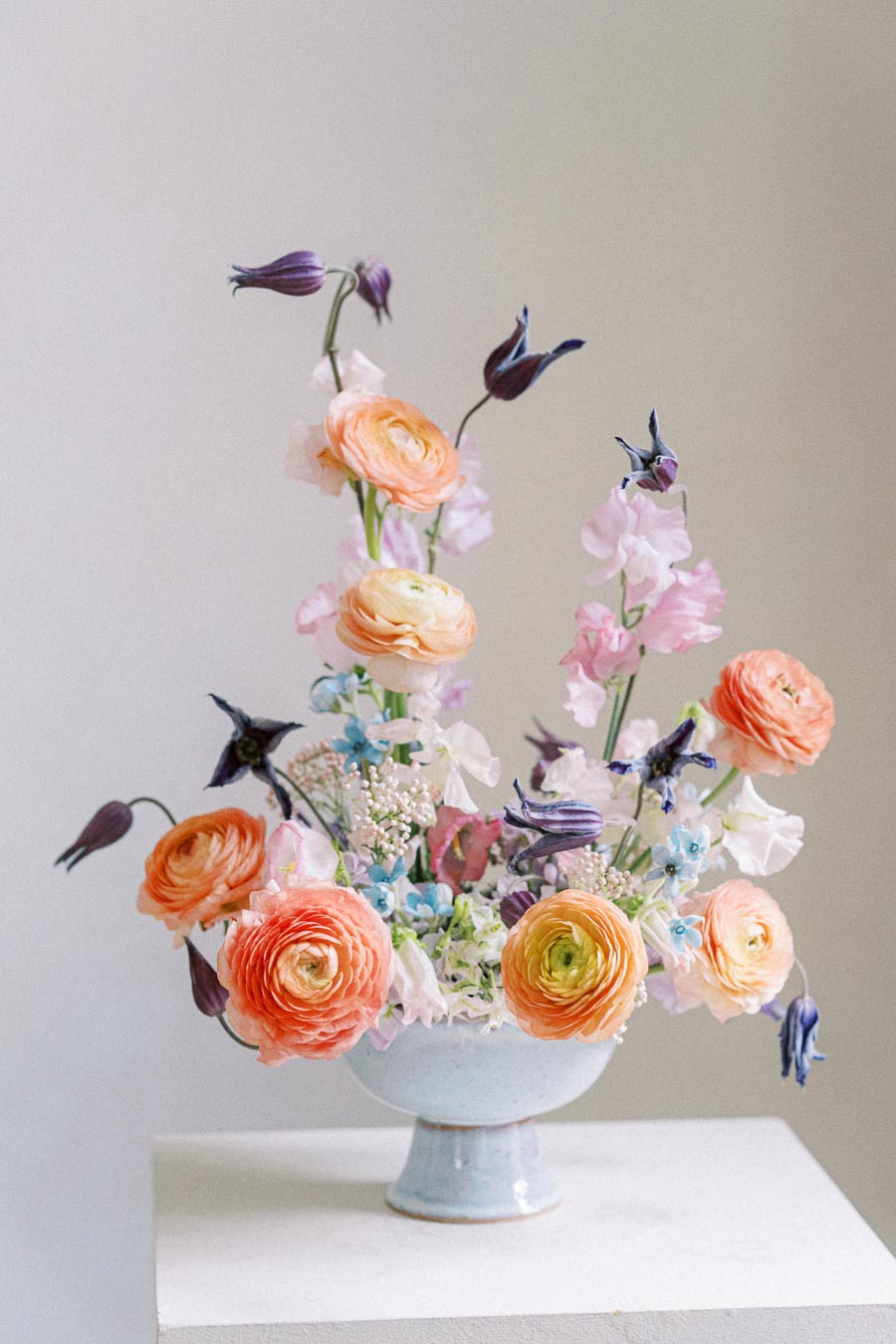 Elegant floral arrangement with orange ranunculus and purple blooms in a white ceramic vase on a pedestal, set against a soft neutral background.