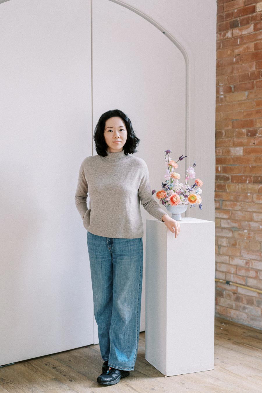 Woman in casual attire standing next to a floral arrangement on a pedestal, in front of a minimalist brick and white background.