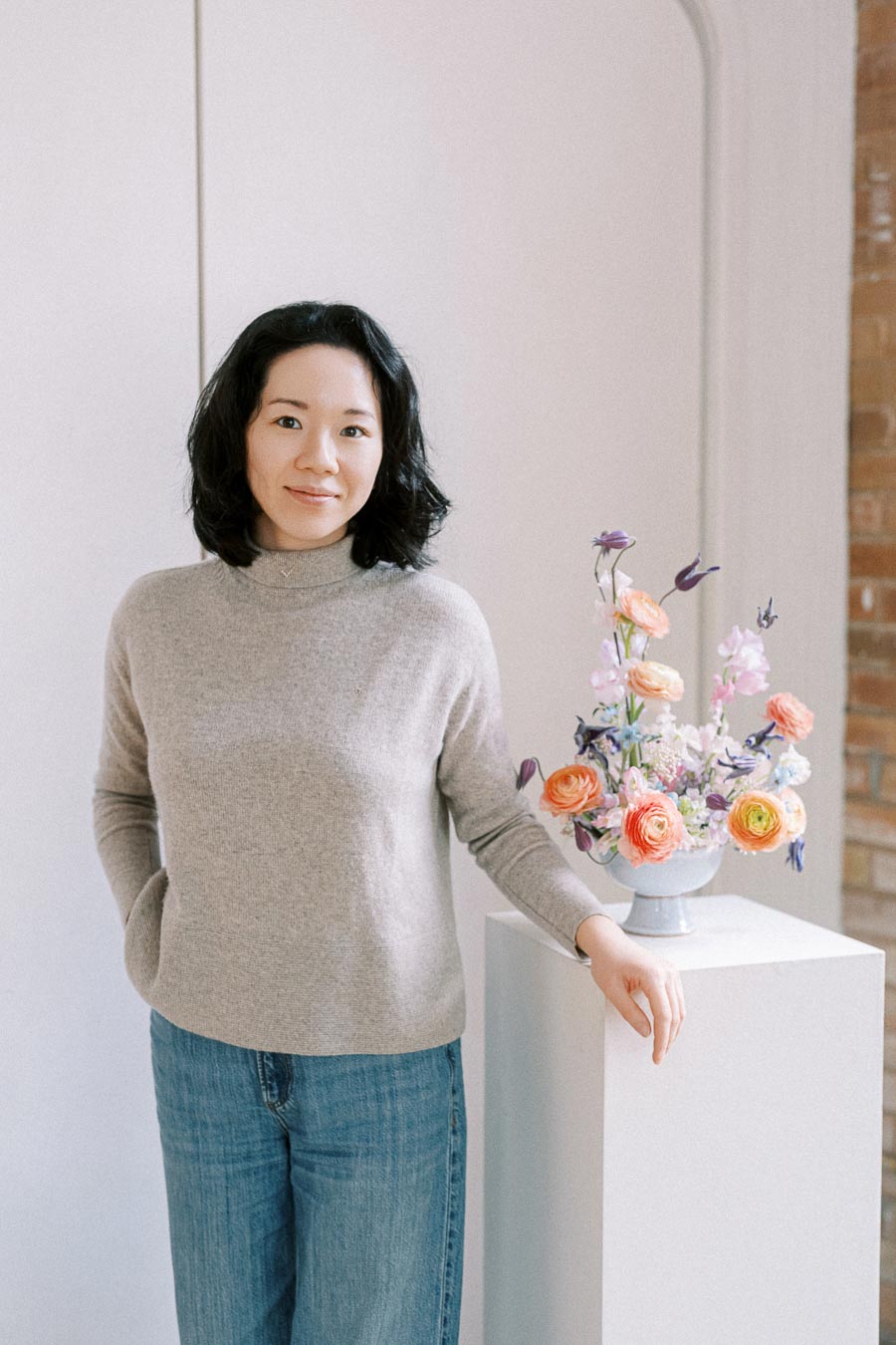 A person wearing a gray sweater and blue jeans standing next to a white pedestal with a colorful flower arrangement in a bright, minimalistic room.