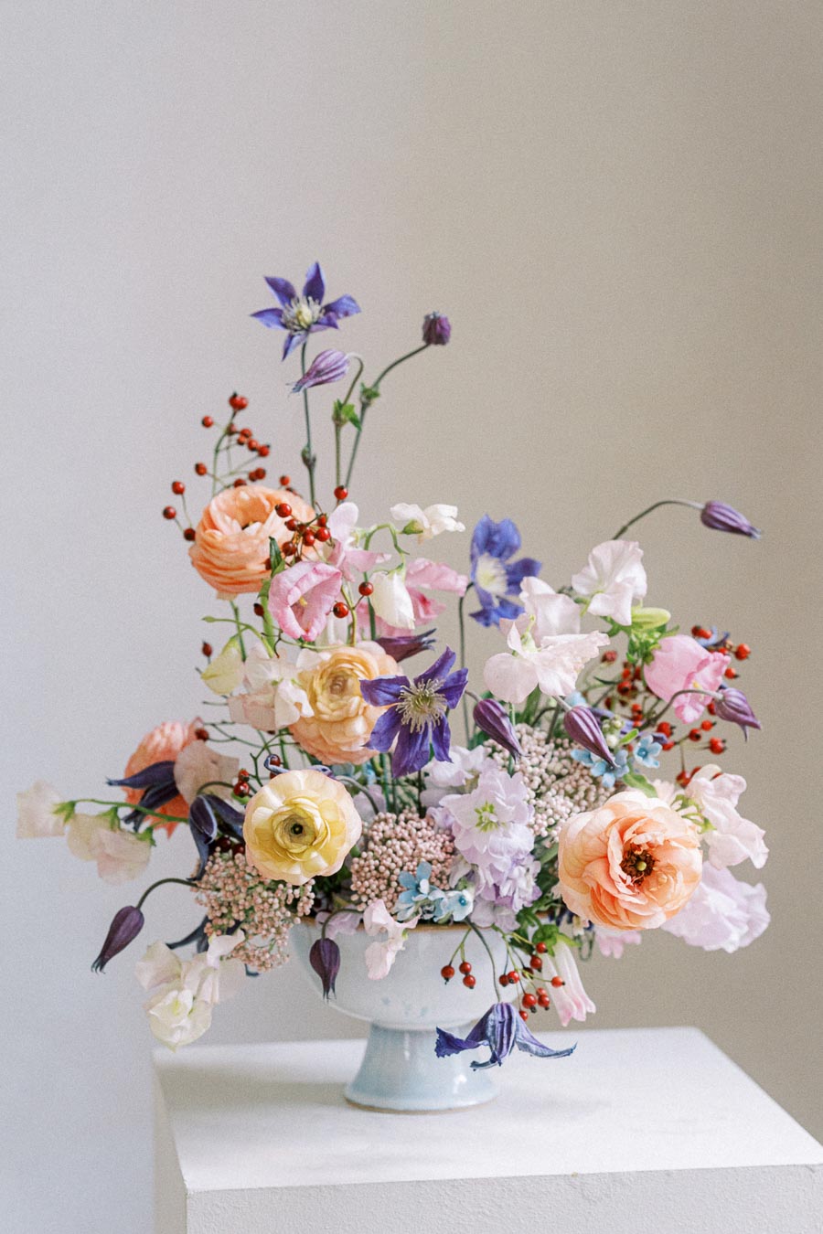 A vibrant floral arrangement featuring a mix of orange, purple, and white flowers with green foliage, artistically displayed in an elegant white vase against a neutral background.