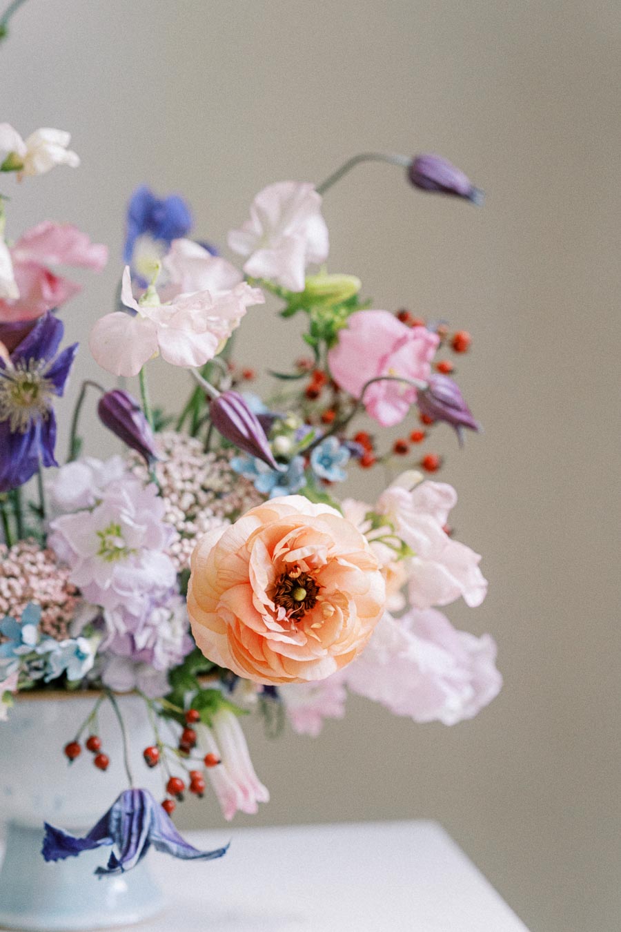 A vibrant floral arrangement featuring an assortment of pastel-colored flowers, including a prominent orange ranunculus, surrounded by pink, purple, and blue blooms, elegantly displayed in a delicate vase.