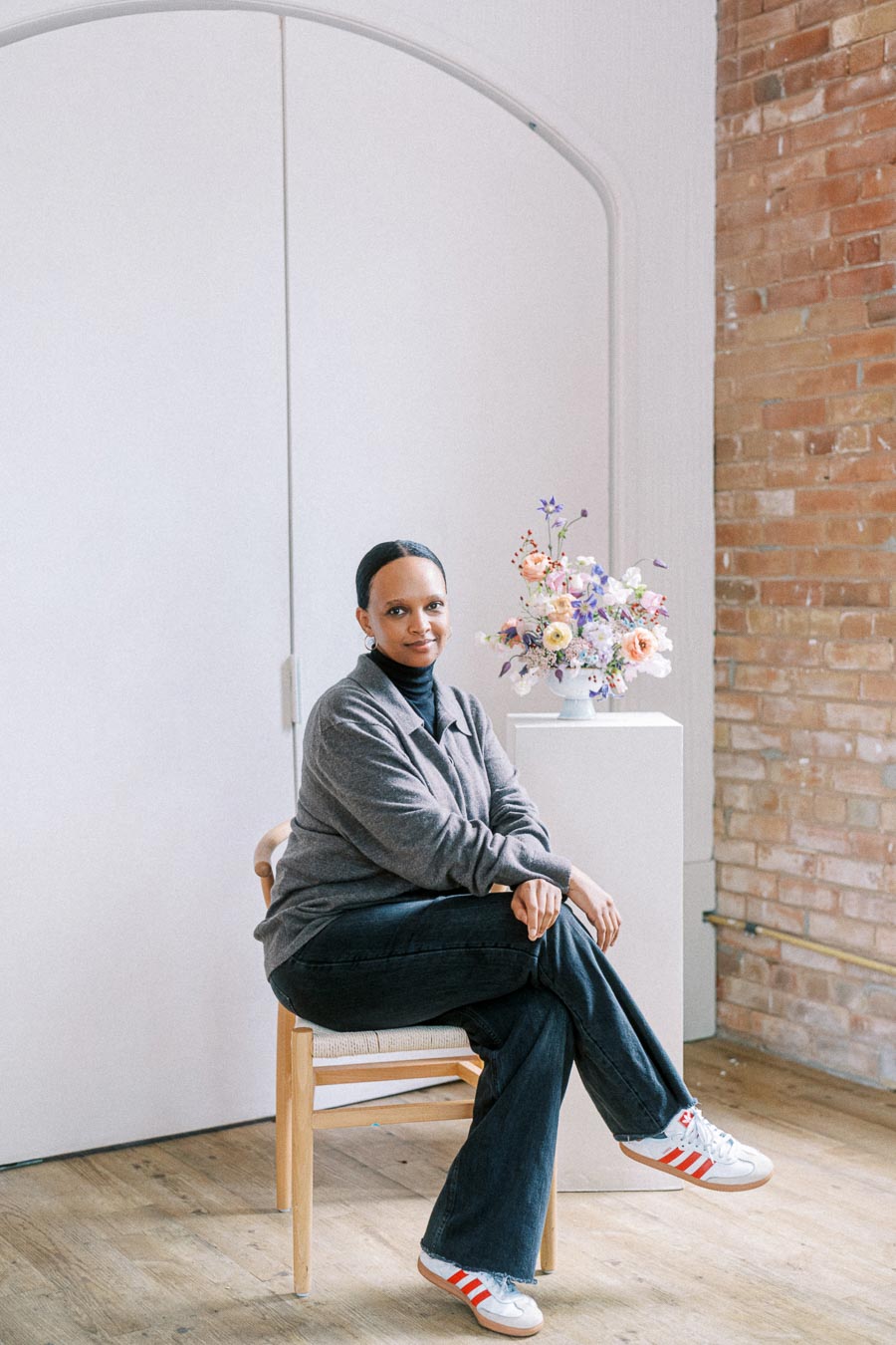 A person sitting on a chair, wearing a gray sweater and jeans, with a vase of colorful flowers on a stand beside them, in a room with a brick wall and wooden floor.