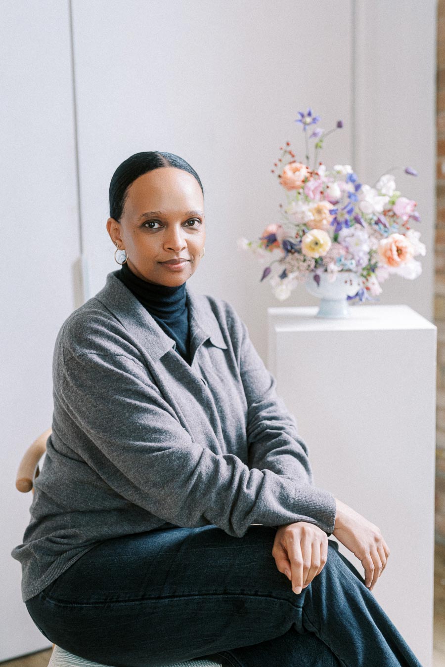 Smiling woman seated indoors next to a vase of colorful flowers, wearing a gray sweater and black turtleneck.