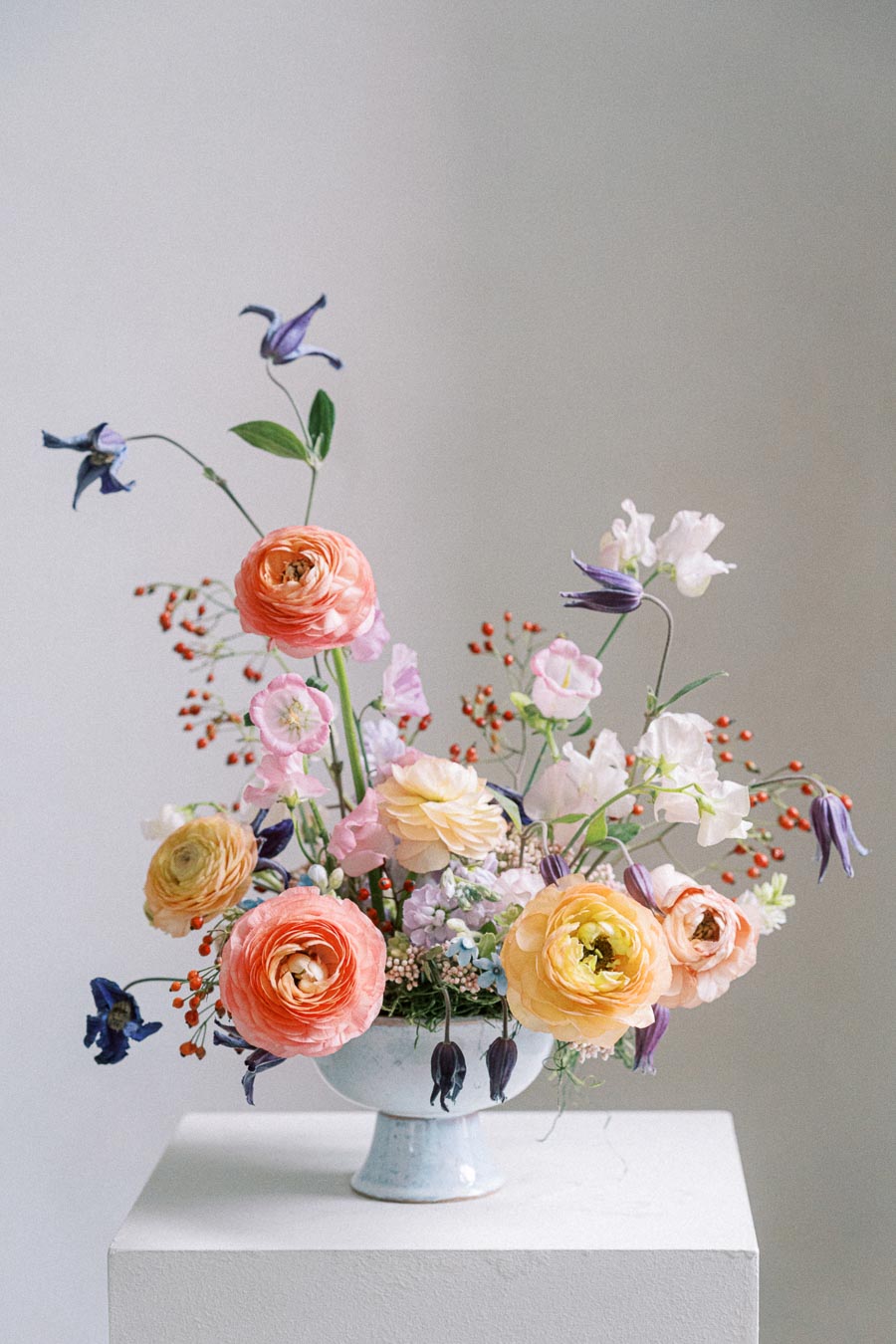 Elegant pastel floral arrangement featuring ranunculus, sweet peas, and clematis in a white ceramic vase on a white pedestal against a neutral background.