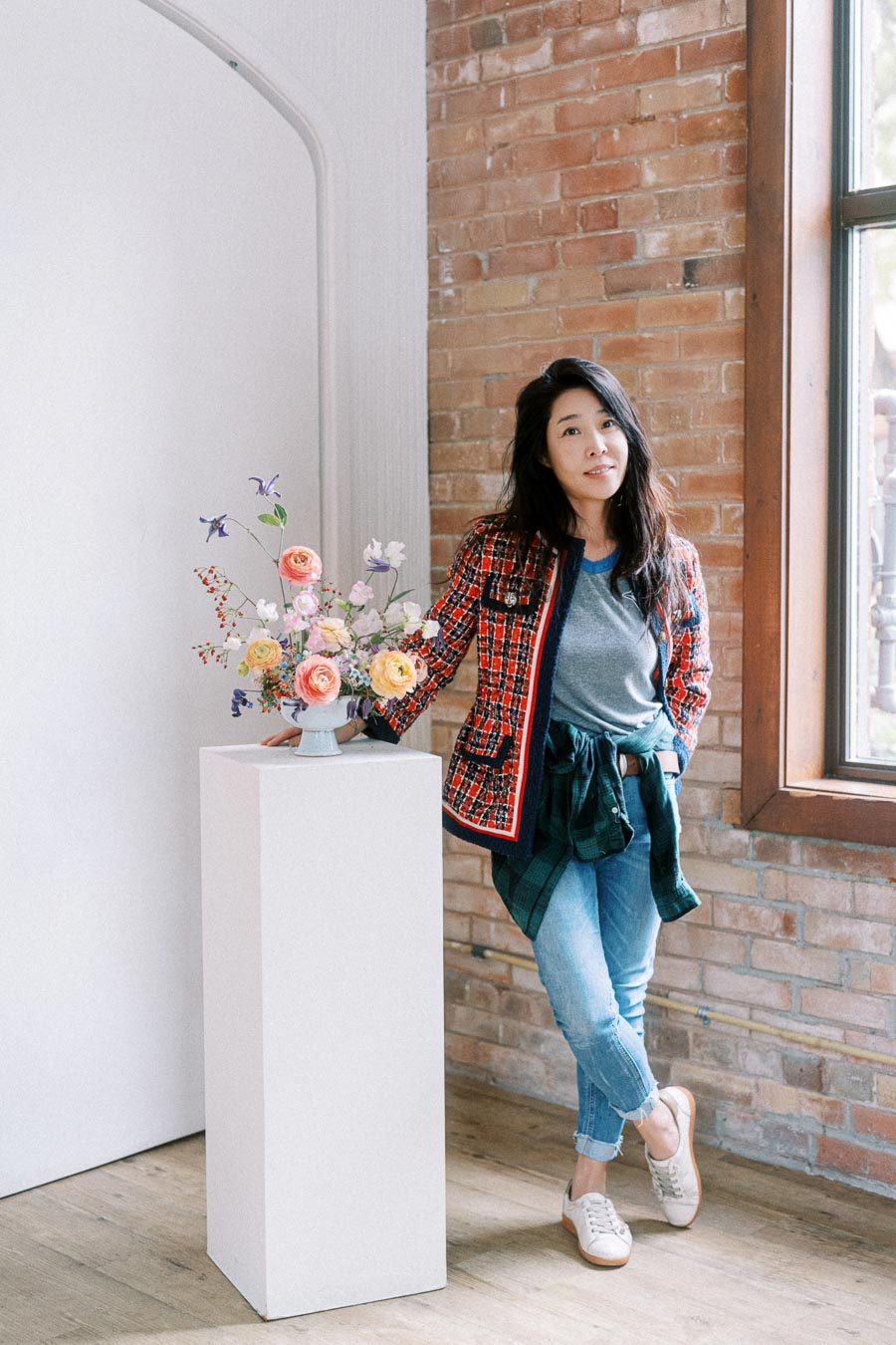 Asian woman in a stylish red and black checkered jacket and jeans, standing by a white pedestal with a floral arrangement, in a bright, modern room with brick walls and large windows.