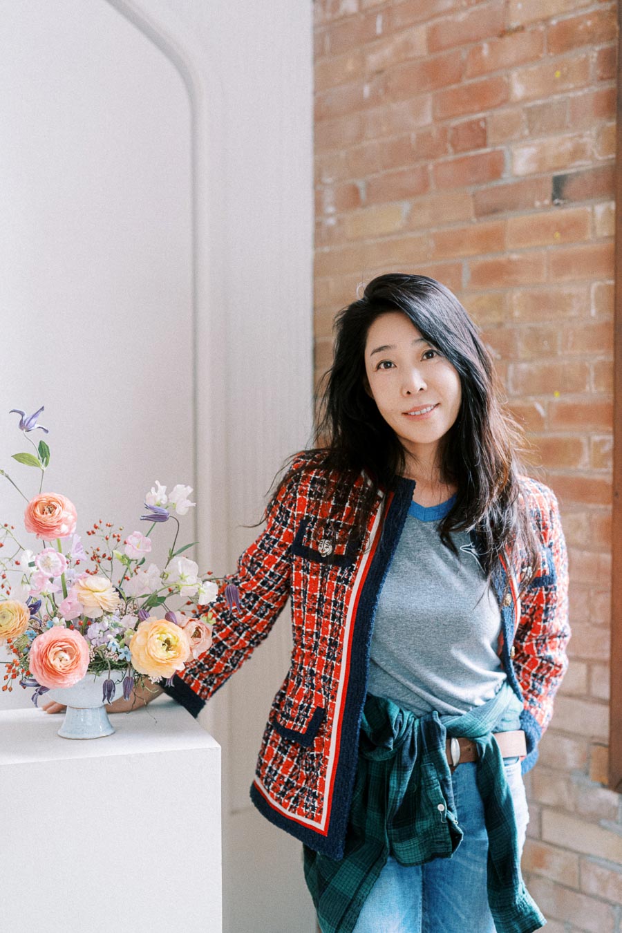 Woman in a stylish checkered jacket and casual attire standing next to a vase of vibrant flowers, against a brick wall background.