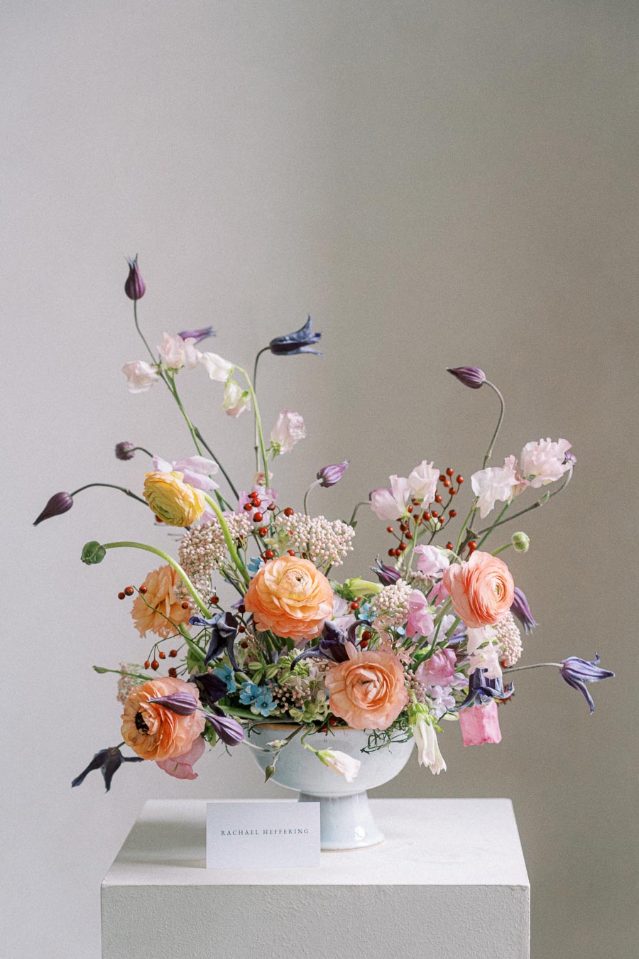 A vibrant floral arrangement featuring orange ranunculus, pink roses, purple bellflowers, and delicate greenery, elegantly displayed in a white ceramic vase against a soft background.