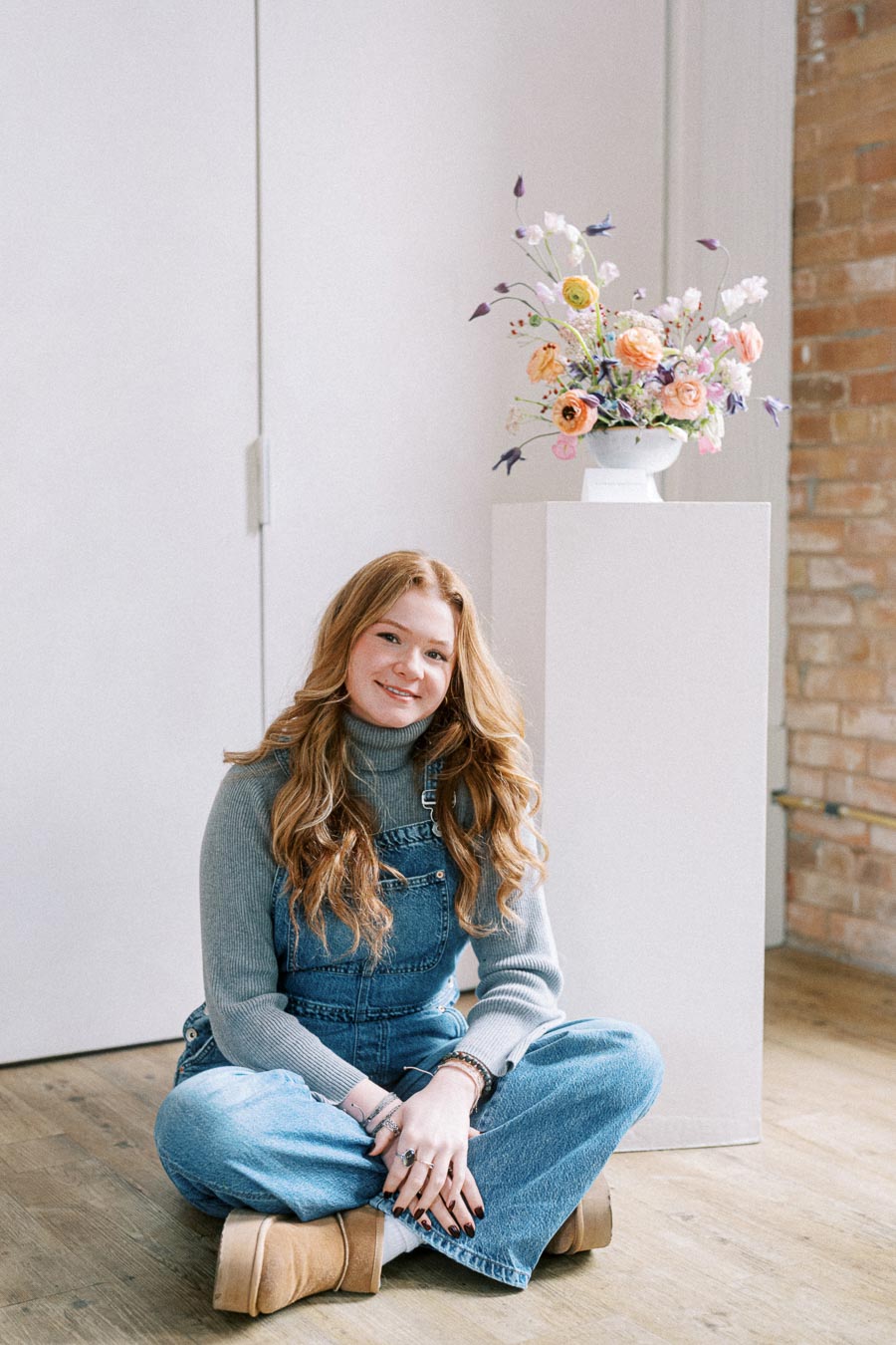 Young woman with long red hair wearing a grey sweater and denim overalls, sitting cross-legged on a wooden floor, smiling in a bright, minimalist room with a flower arrangement on a pedestal.