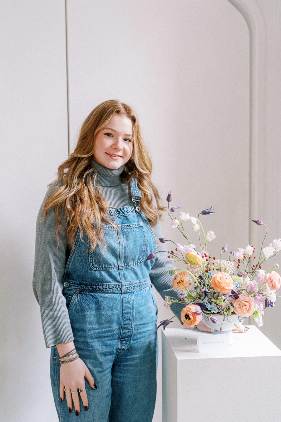 Young woman in denim overalls and gray sweater standing beside a colorful floral arrangement in a bright, minimalistic room.