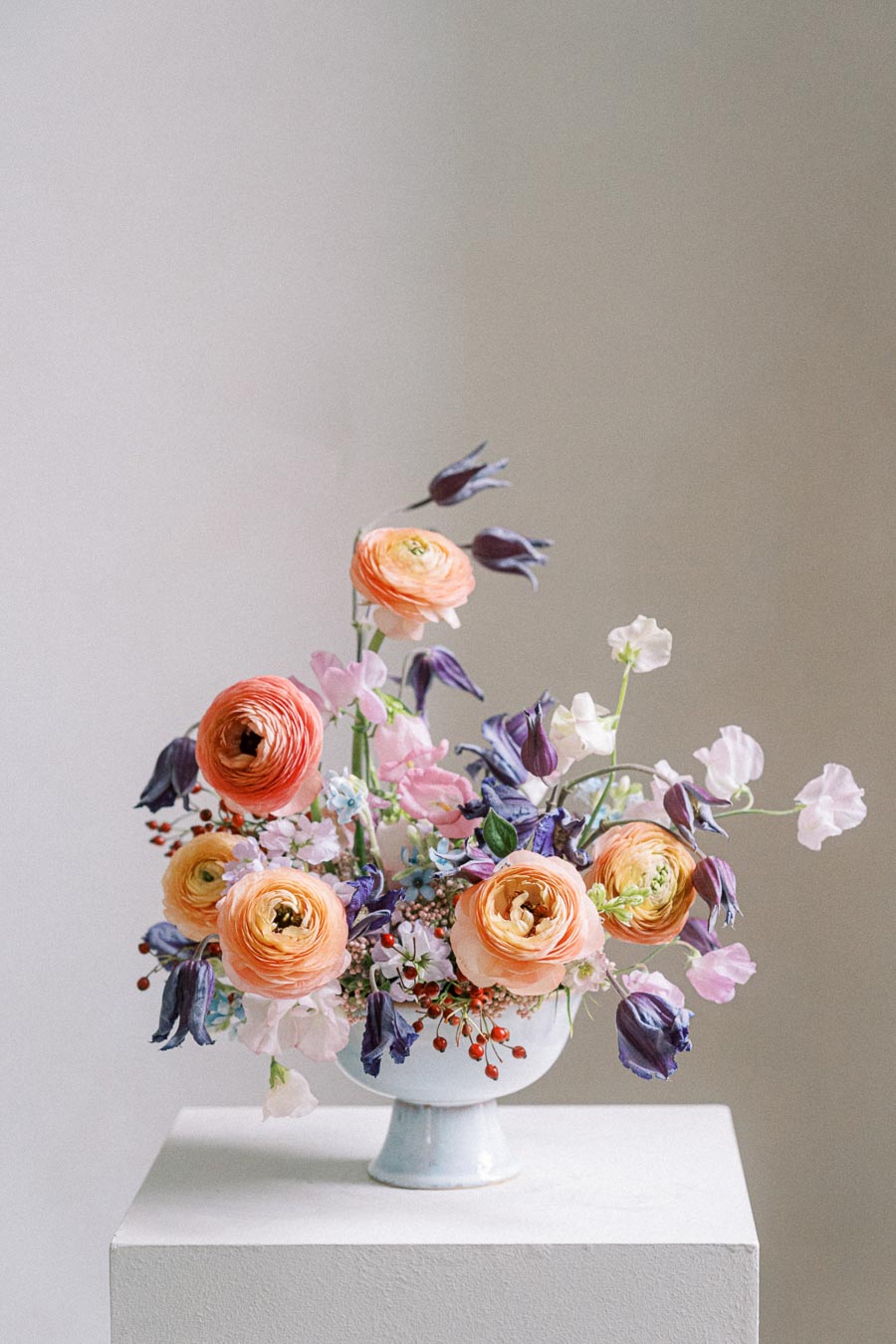 Elegant floral arrangement with vibrant orange, pink, and purple flowers in a white vase on a pedestal, against a neutral background.