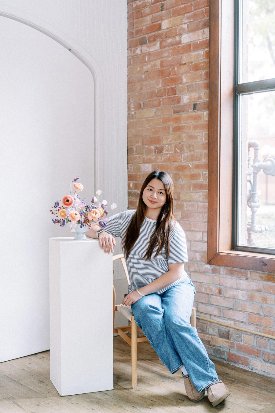 Woman sitting on a chair beside a pedestal with a colorful flower arrangement, in a bright room with a brick wall and window. Casual attire and serene ambiance.