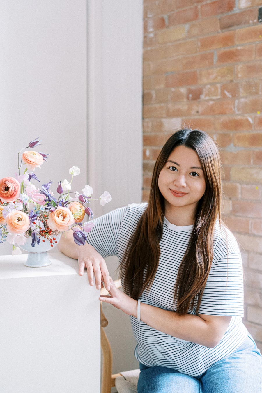 A woman in a striped shirt sits at a white table displaying a vibrant floral arrangement with peach and purple flowers, set against a warm, brick wall background.