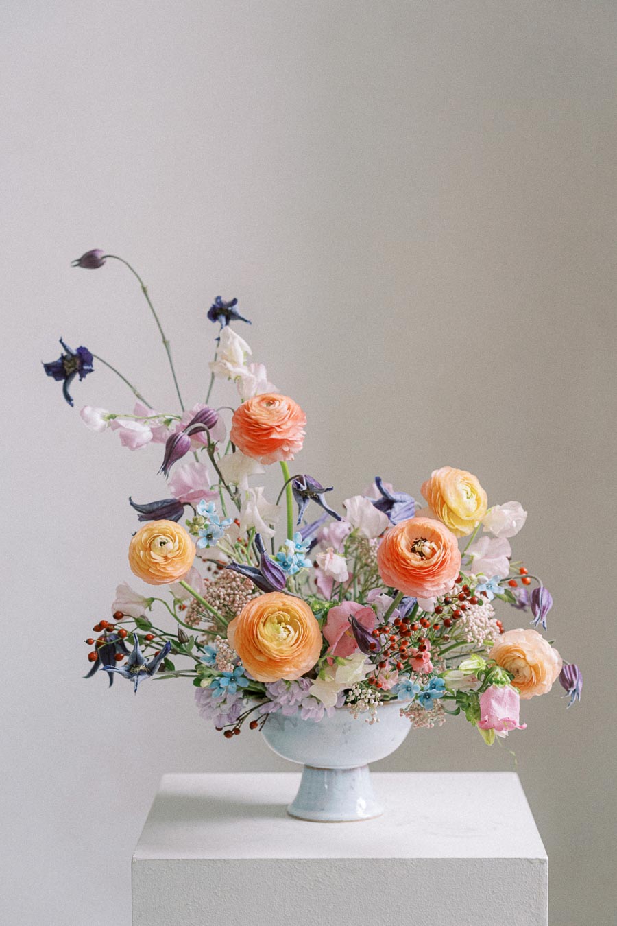 Elegant floral arrangement featuring orange and yellow ranunculus, purple clematis, and assorted blooms in a white pedestal vase, set against a neutral background