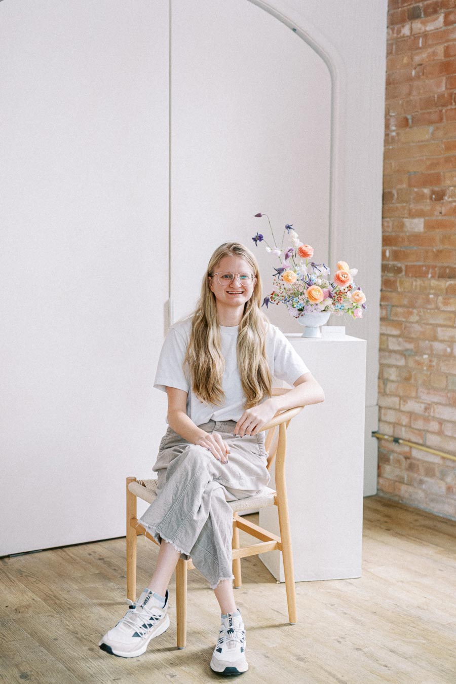 Young woman seated in a modern, minimalist room with exposed brick wall and wooden flooring, next to a table with a colorful floral arrangement.