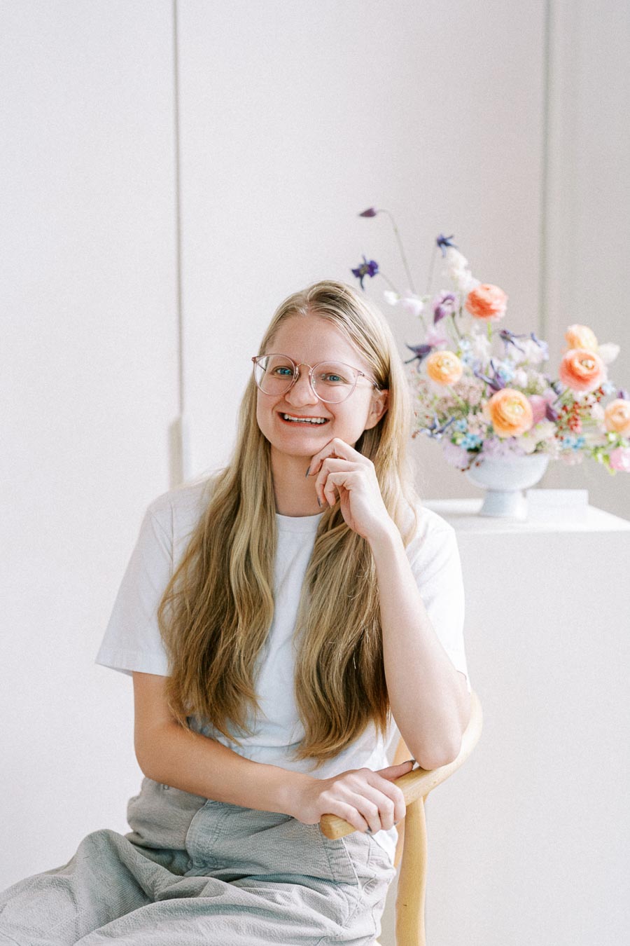 Young woman with long blonde hair and glasses seated beside a colorful flower arrangement in a bright, minimalist interior.