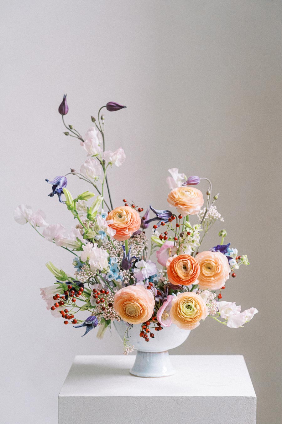 Beautiful floral arrangement featuring peach ranunculus, pink sweet peas, and purple clematis in a white vase on a pedestal against a neutral background.