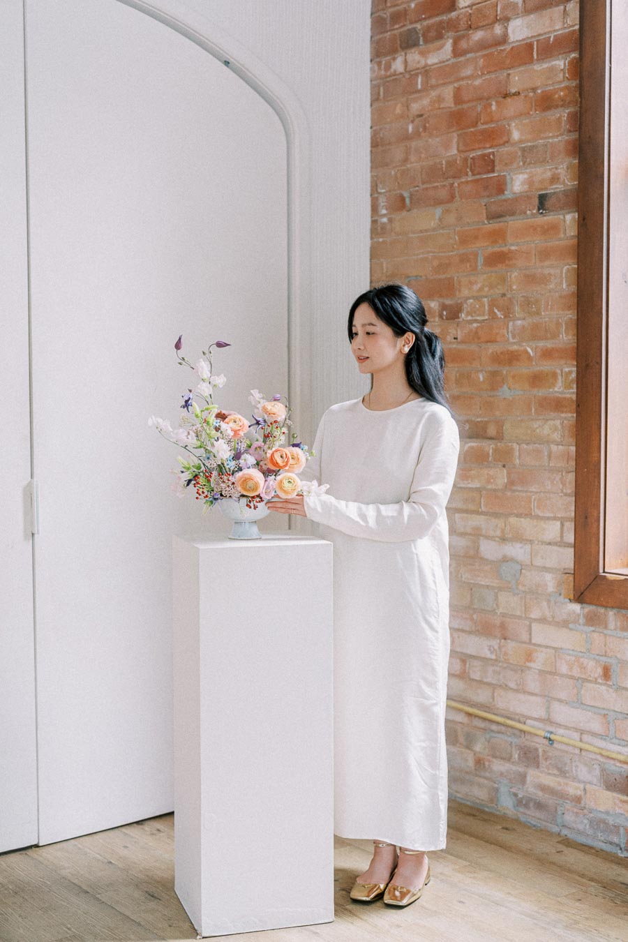 A person in a white dress arranging a colorful flower bouquet on a pedestal inside a room with a brick wall and wooden floor.