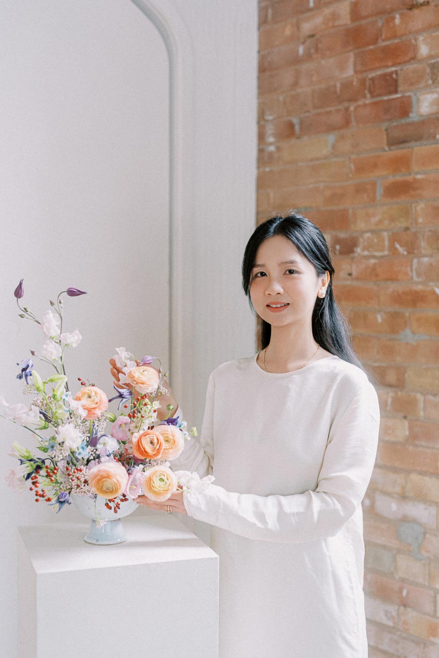 A woman in a white dress stands beside a vibrant floral arrangement in a vase, with a brick wall in the background.