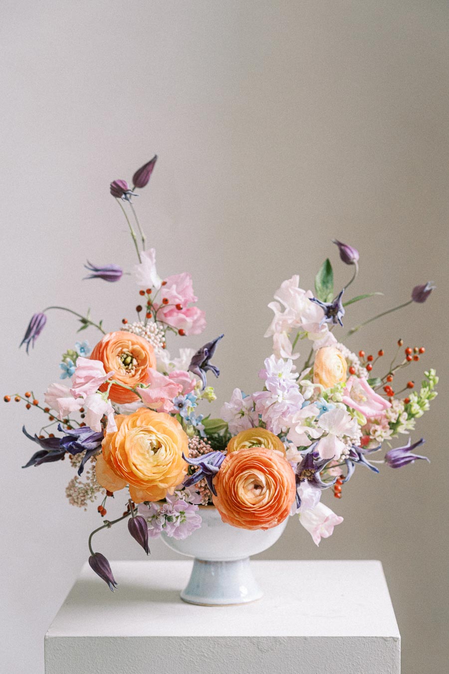 Elegant floral arrangement in a white vase featuring vibrant orange ranunculus, delicate pink blossoms, purple accents, and lush greenery, displayed on a white pedestal against a neutral background.