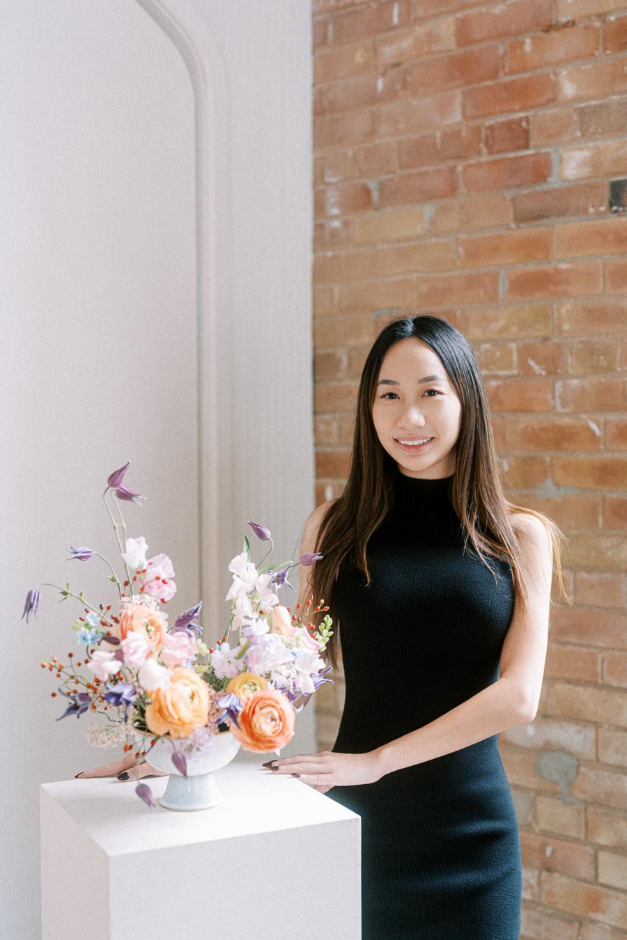 A woman in a black dress standing next to a white pedestal with a vase of colorful flowers, set against a textured brick wall backdrop.