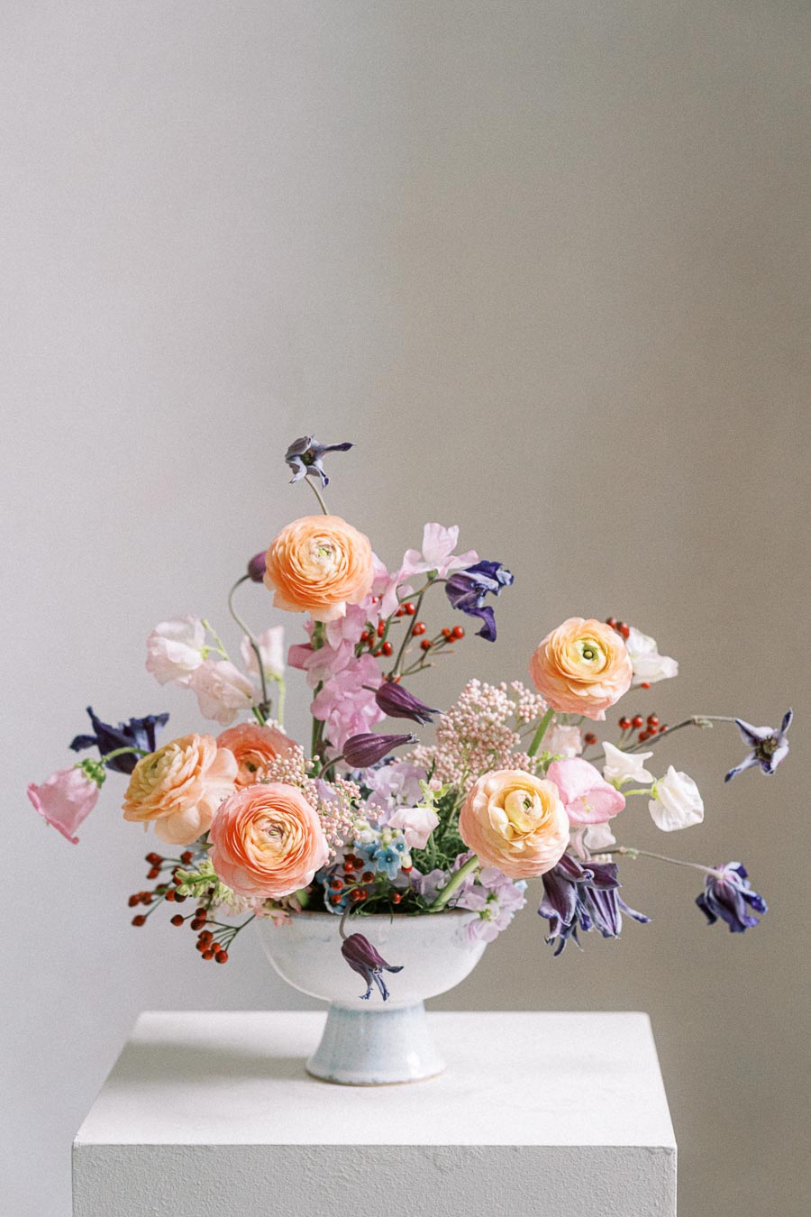 A vibrant floral arrangement in a white ceramic vase featuring peach ranunculus, purple bellflowers, pink sweet peas, and blue accents on a white pedestal against a neutral background.