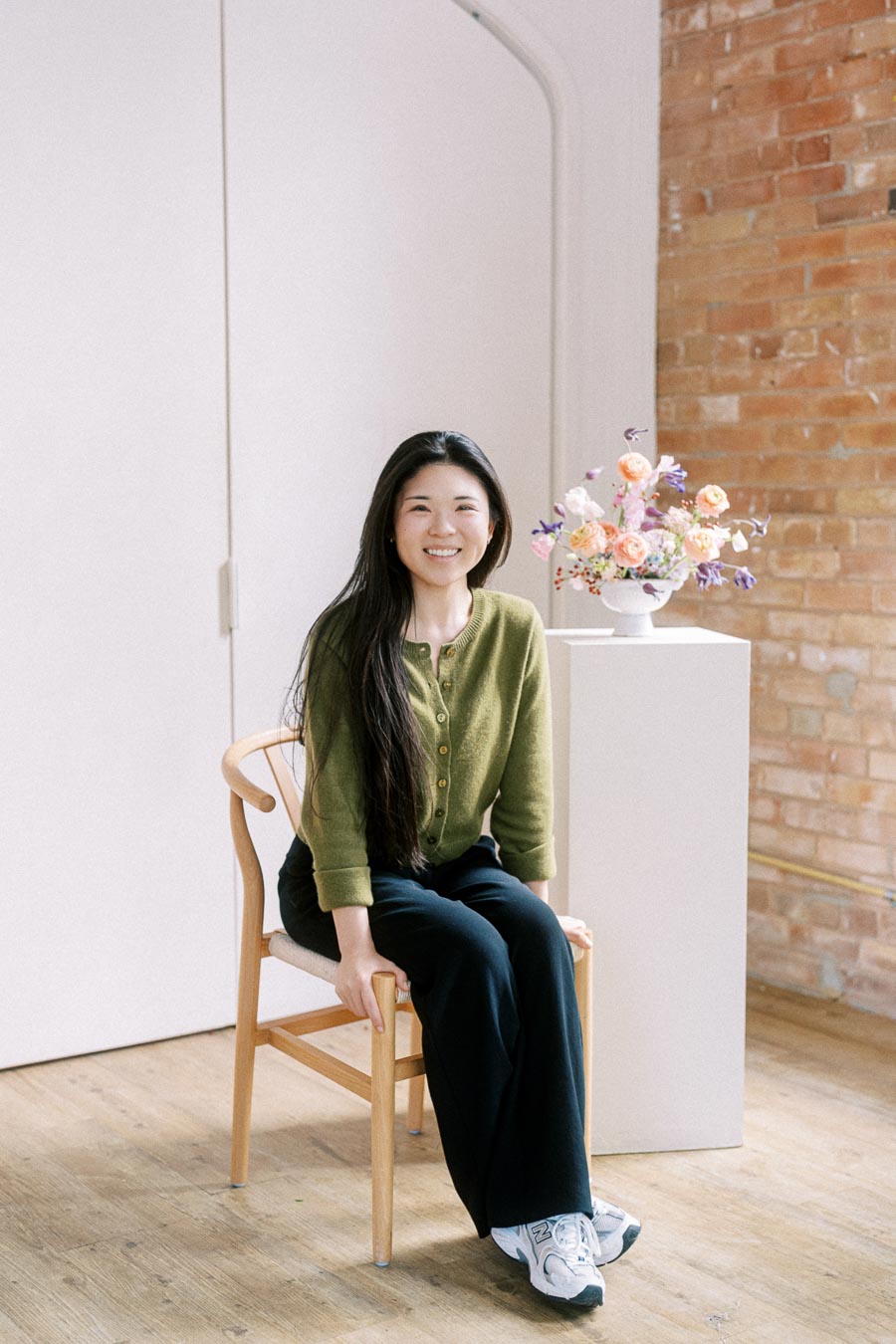 A person sitting on a wooden chair in a well-lit room with a brick wall, wearing a green sweater and black pants, smiling at the camera. Next to them is a white pedestal displaying a colorful floral arrangement.