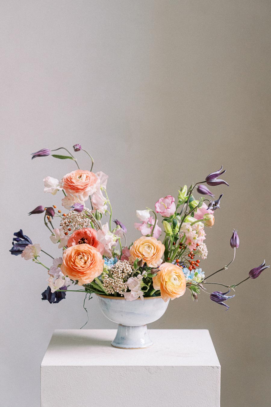 Elegant floral arrangement with orange and yellow roses, purple buds, and mixed greenery in a white vase, placed on a pedestal against a neutral background. Perfect for wedding or event decor inspiration.