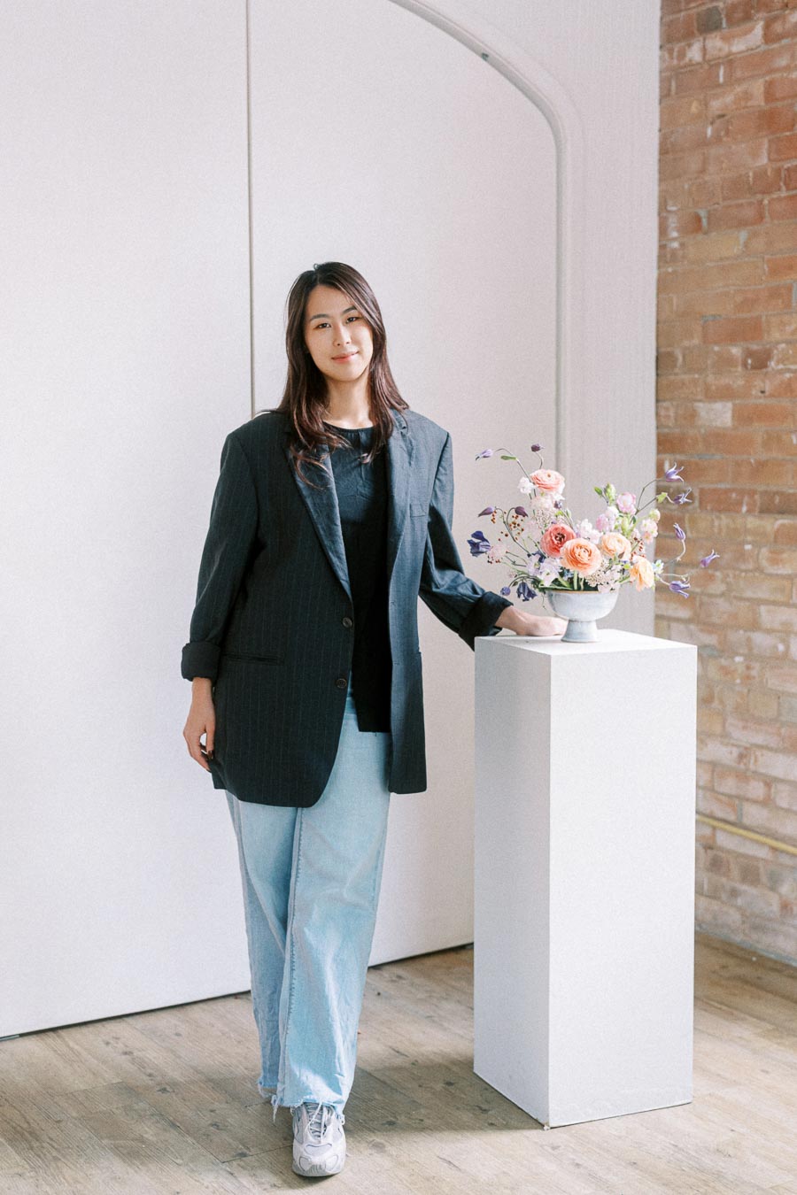 Woman in a black blazer and light jeans standing next to a floral arrangement on a pedestal, in a minimalist room with brick and white walls.