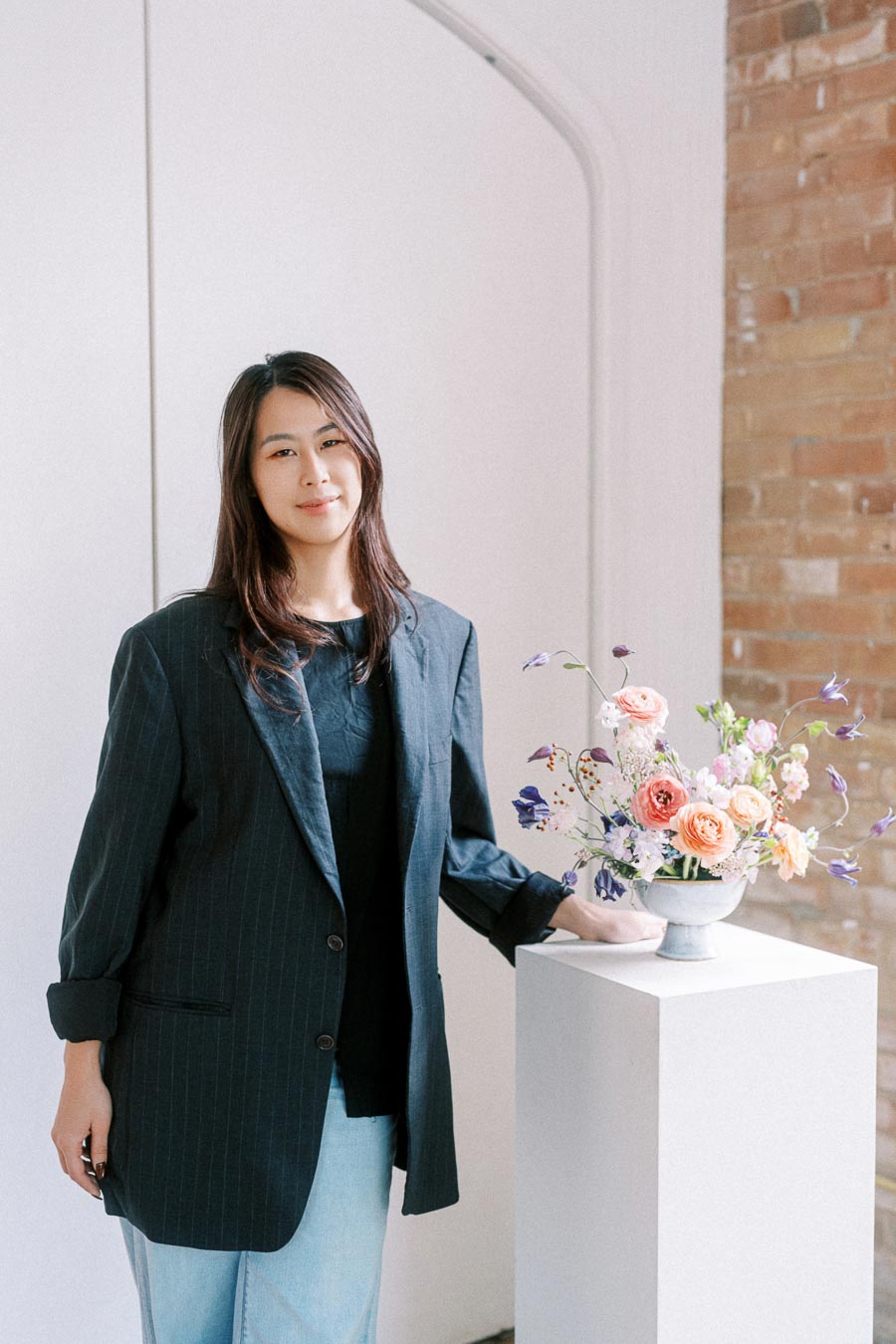 A person in a dark blazer and jeans stands beside a pedestal with a colorful floral arrangement.