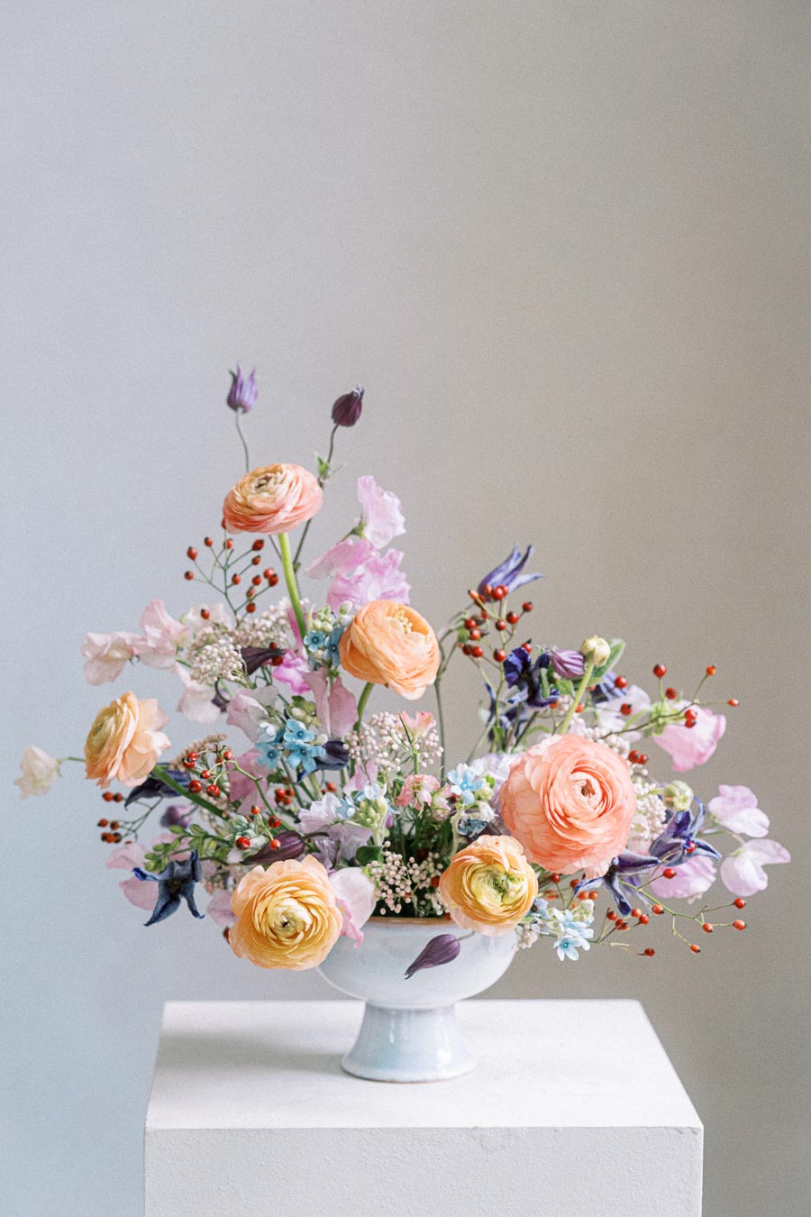 A vibrant floral arrangement in a white ceramic vase featuring peach and pink ranunculus, delicate purple and blue blossoms, and red berry accents, set against a neutral background. Ideal for wedding decor or spring centerpiece inspiration.