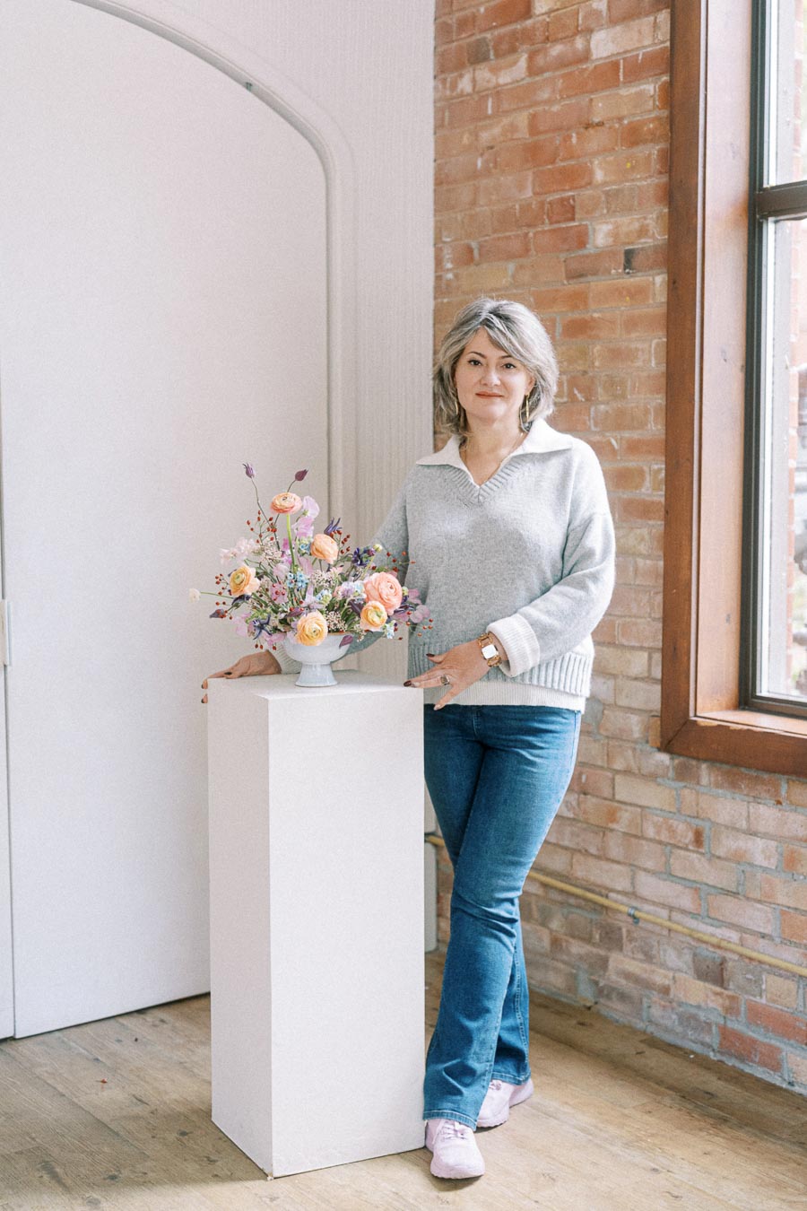 A woman standing next to a floral arrangement on a white pedestal, wearing a gray sweater and jeans, in a room with exposed brick walls and a large window.