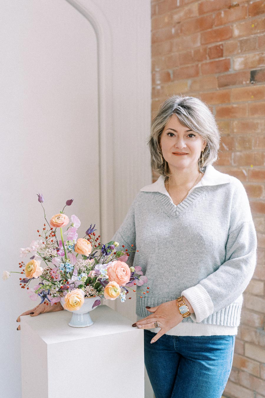 A woman with a smiling expression standing beside a floral arrangement in a vase, set against a brick wall background. She is wearing a light gray sweater and blue jeans, highlighting a casual yet elegant style. The flowers are a mix of pastel colors, adding a touch of vibrancy to the scene