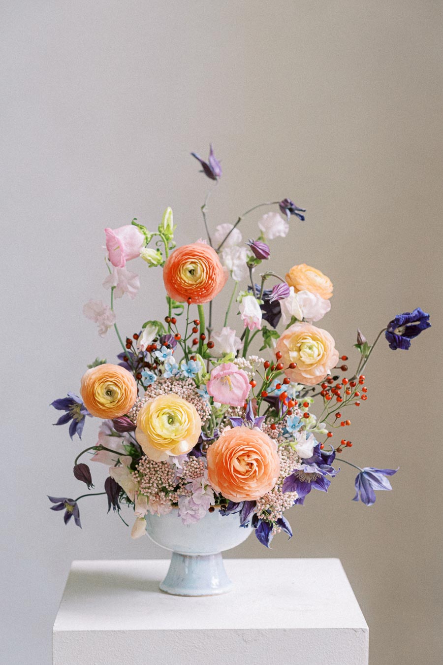 A vibrant floral arrangement featuring colorful ranunculus, sweet peas, and various wildflowers in a white ceramic vase on a minimalist pedestal, set against a neutral background.