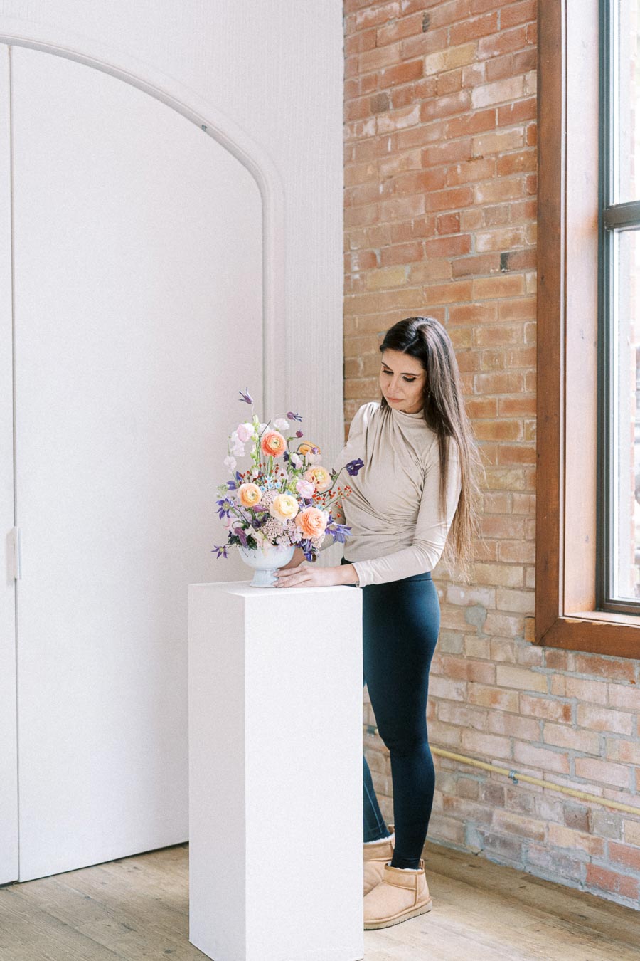 A woman arranging a colorful flower bouquet in a vase on a pedestal against a brick wall background in a bright room.