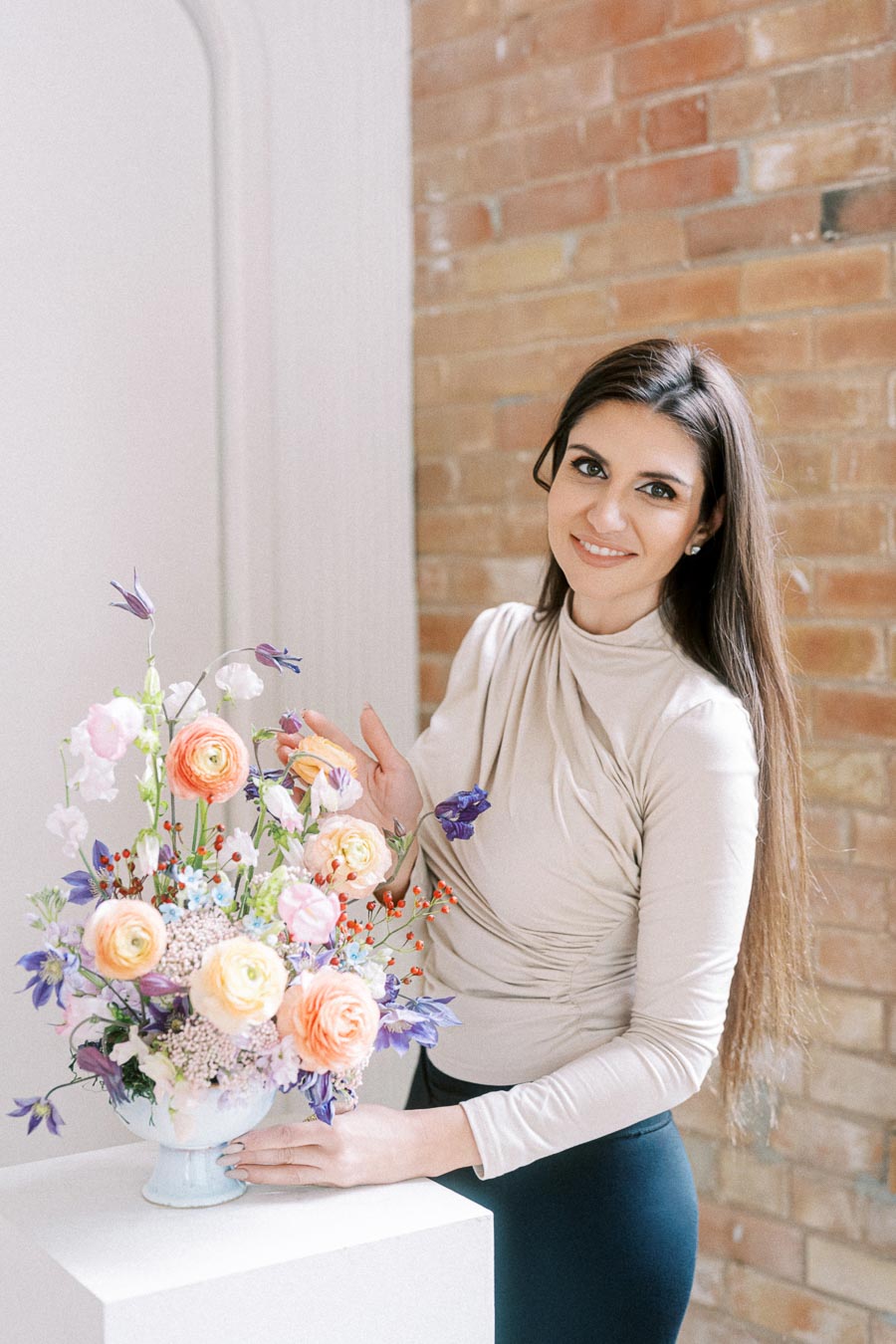 A woman in a beige top smiling next to a colorful floral arrangement in a decorative vase, set against a rustic brick wall.