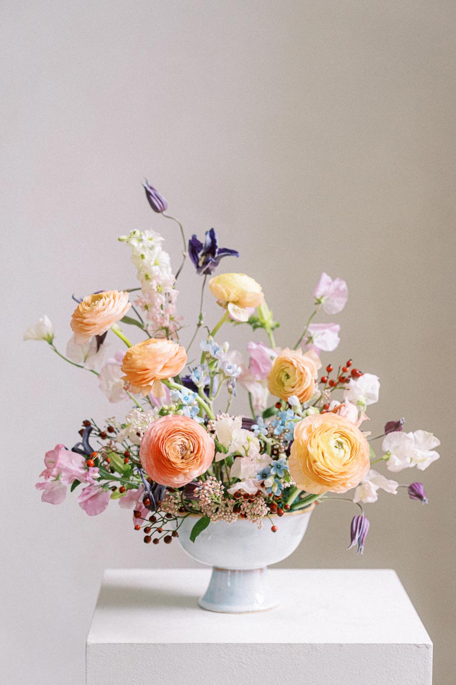 Elegant floral arrangement in a white vase featuring vibrant orange and yellow ranunculus, purple and pink blossoms, and lush greenery, perfect for weddings or home decor inspiration.