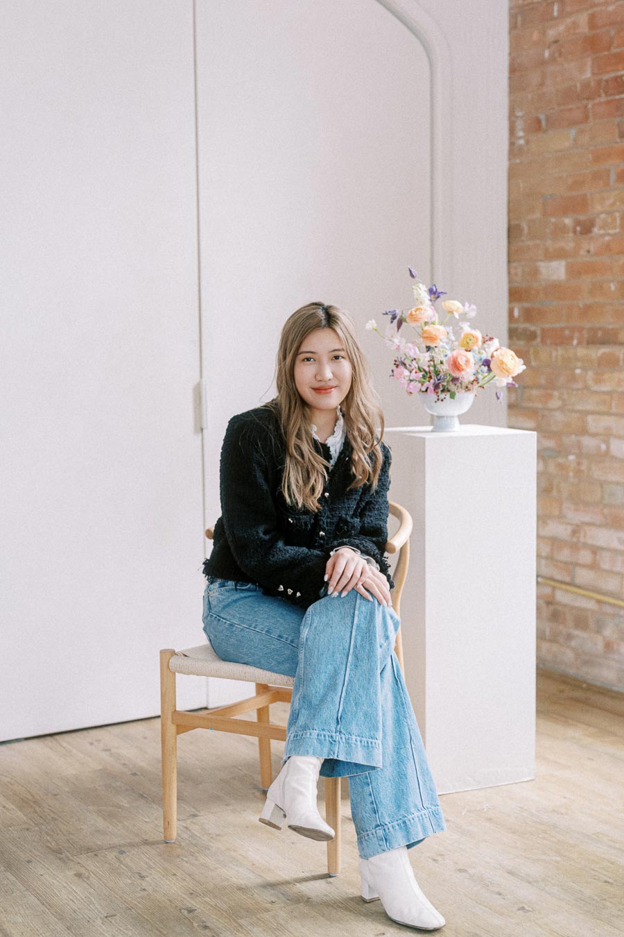 A person sitting on a wooden chair in a modern, minimalist room with wooden flooring and exposed brick wall. They are wearing a black sweater, blue jeans, and white ankle boots. A stylish floral arrangement in a white vase is on a pedestal beside them.