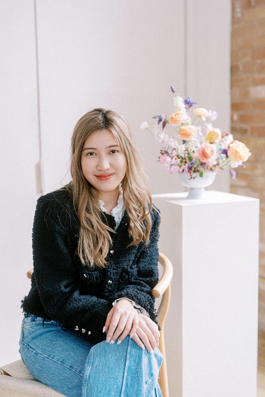 A woman with long, light brown hair sitting in a chair, wearing a black jacket and jeans, with a vase of colorful flowers on a pedestal in the background. The setting is a softly lit, modern interior with white walls and a touched of brick.