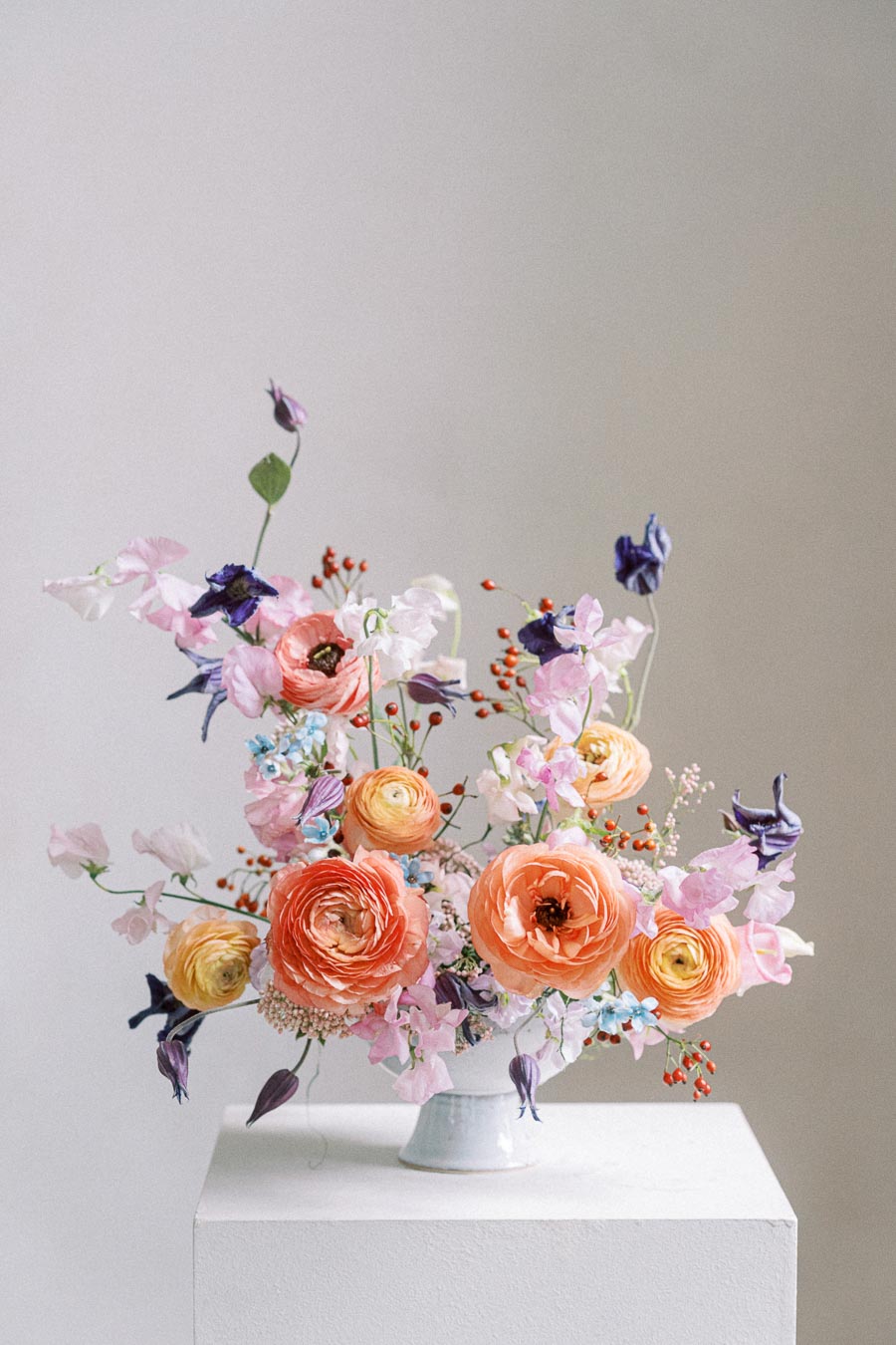 A vibrant flower arrangement featuring orange, pink, and yellow blooms with purple and blue accents in a white vase, set against a neutral background.