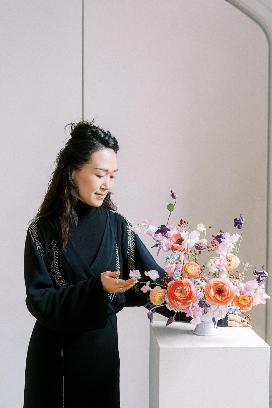 A person arranging a colorful floral bouquet in a white vase on a pedestal, wearing a black outfit with delicate embroidery, in a minimalist indoor setting.