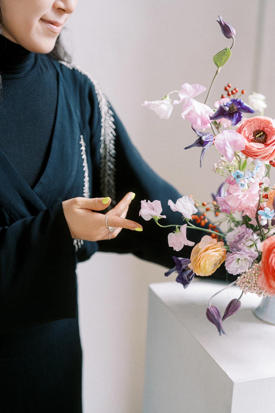 A person in a black dress arranging a colorful floral bouquet with various flowers, including pink, purple, and orange blooms, on a white pedestal.