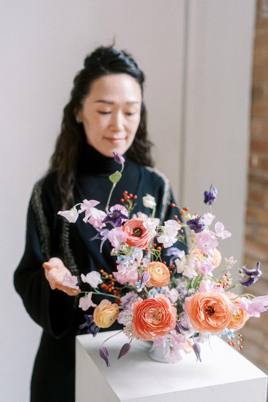 A person arranging a vibrant floral bouquet with orange, pink, and purple flowers on a white pedestal, showcasing delicate flower arranging skills in a bright room.