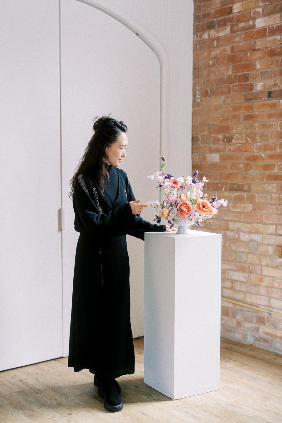 A person arranging a vibrant flower bouquet on a pedestal against a stylish brick wall, showcasing floral design skills in an elegant setting.