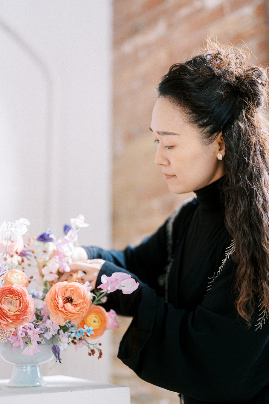 A person arranging a colorful bouquet of flowers in a vase, featuring orange and pink blooms with delicate petals, set against a softly blurred indoor background.