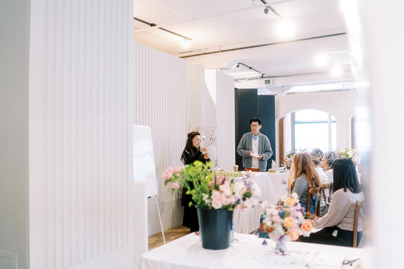 A group of people attending a floral workshop in a modern, well-lit room, with two presenters standing near a table filled with vibrant flower arrangements, engaging with the audience seated around them.